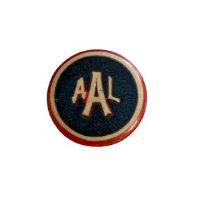 May include: A circular button featuring a dark blue textured center with the letters AAL printed in a cream color. The button has a distinct red outer border and a thin cream-colored ring separating the red edge from the dark blue center area.