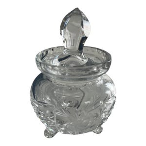 Pode incluir: Um frasco de vidro transparente decorativo com tampa. O frasco tem um corpo arredondado com padrões florais gravados e assenta em três pequenos pés. A tampa apresenta um remate pontiagudo. O frasco foi concebido para guardar pequenos objetos.