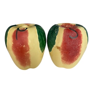 May include: A pair of ceramic apple-shaped salt and pepper shakers. The shakers are painted with a yellow base, a red stripe, and green leaves. These apple shakers are a decorative kitchen item.
