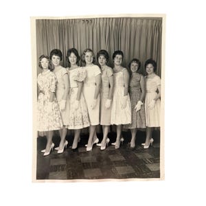May include: A black and white vintage photograph featuring eight individuals standing in a row against a curtain backdrop. Each person wears a light-colored dress, white gloves, and high-heeled shoes. The dresses vary in style, including floral patterns, solid colors, and different sleeve lengths, all reflecting mid-century fashion.