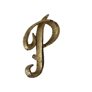 May include: A gold-toned metal brooch shaped like the capital letter P. The surface of the letter features a textured, brushed finish with visible vertical lines throughout the design. The script-style letter has a curved, elegant form.