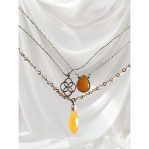 Peut inclure: Trois colliers superposés avec des chaînes argentées. Le collier supérieur a un pendentif argenté avec un motif à quatre pétales. Le collier du milieu a un pendentif orange en forme de goutte. Le collier inférieur a un long pendentif jaune facetté.