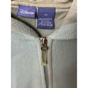 May include: Close-up of a light blue Disney fleece hoodie with a zipper. The hoodie has a gray hood and a tag that reads "Disney" and "M". The tag also states the material is 100% polyester and made in Macau.