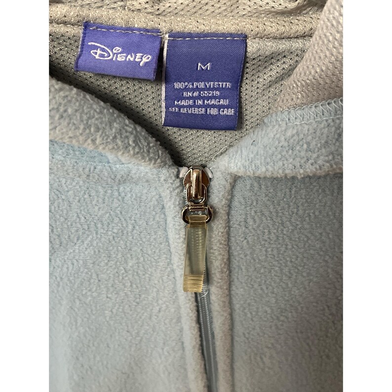 May include: Close-up of a light blue Disney fleece hoodie with a zipper. The hoodie has a gray hood and a tag that reads "Disney" and "M". The tag also states the material is 100% polyester and made in Macau.