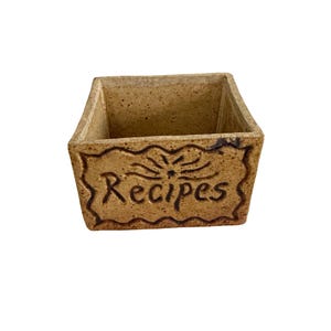 May include: A square, ceramic recipe box in a speckled tan color. The word "Recipes" is carved into a decorative border on the front. The box has a simple, rustic design.