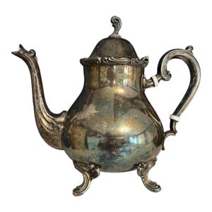Puede incluir: Tetera plateada con un pico y asa curvados. La tetera tiene un cuerpo redondeado, una tapa con un adorno decorativo y descansa sobre tres patas ornamentadas. La superficie metálica refleja la luz, mostrando una estética vintage.