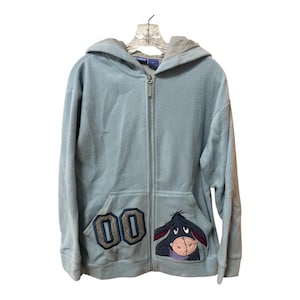 May include: A light blue zip-up hoodie with a gray hood lining. The front pocket features the number "00" and an embroidered image of Eeyore from Winnie the Pooh. The hoodie is hanging on a clear plastic hanger.