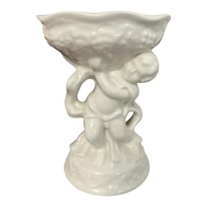 Puede incluir: Un cuenco de pedestal de cerámica blanca con una figura de querubín que sostiene el cuenco. El cuenco tiene un borde con volantes y el querubín está en una pose clásica. La base del pedestal tiene un diseño floral decorativo.
