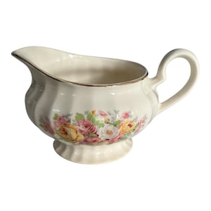 Peut inclure: Une saucière crème avec un motif floral de roses roses, jaunes et blanches. Elle possède un bord doré et une anse incurvée. La base est évasée et le bec verseur est légèrement courbé. Idéale pour servir des sauces.