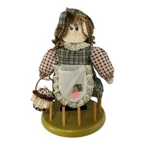 Puede incluir: Muñeca decorativa con vestido de cuadros verdes, delantal blanco y gorro a juego. La muñeca tiene el pelo castaño rizado y sostiene una pequeña cesta. Está montada sobre una base de madera con clavijas de madera.