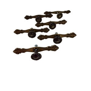 May include: A set of five antique brass-toned metal drawer pulls. Each pull features an elongated, curved horizontal bar with decorative scrollwork at the ends and a central circular knob with a scalloped edge and a visible mounting screw.