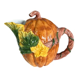May include: A ceramic pumpkin-shaped creamer with a brown handle and spout. The creamer is orange with green, yellow, and brown leaf and vine accents. The interior of the creamer is white.