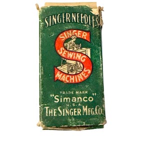 May include: A rectangular green cardboard box for Singer needles. The front features a large red letter S with the text Singer Sewing Machines, an illustration of a person at a sewing machine, and the text Simanco U.S.A. The Singer Mfg. Co.