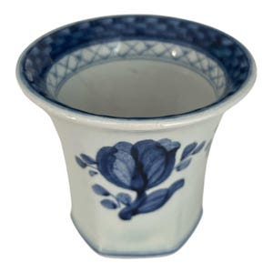 Puede incluir: Un jarrón de cerámica blanco con un diseño floral azul. El jarrón tiene un borde acampanado con un patrón geométrico azul y blanco. El cuerpo del jarrón presenta un diseño de tulipán azul.