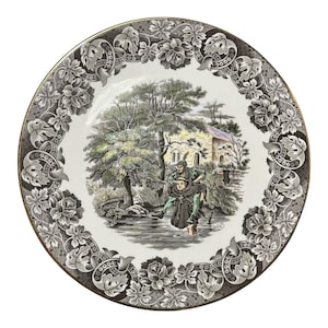 May include: Round decorative plate with a detailed black and white floral border and a gold rim. The center features a pastoral scene with figures in a forest and a building in the background. The plate is white.