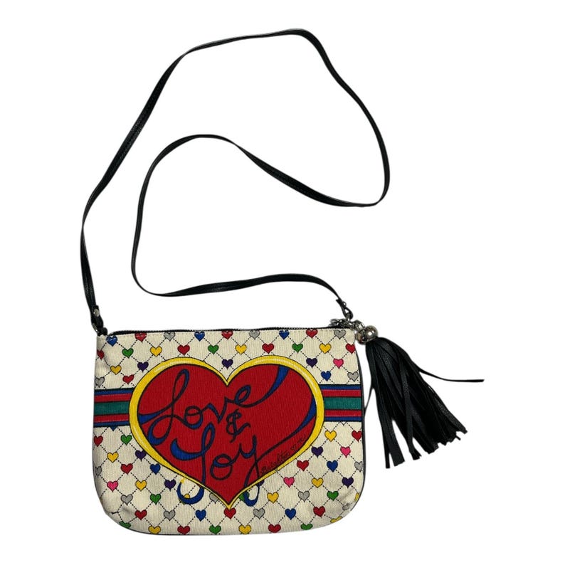 Op de afbeelding: Cr&egrave;mekleurige crossbodytas met een zwarte band en kwast. De tas heeft een groot rood hart met de woorden "Love & Joy" in cursief, omgeven door een kleurrijk hartpatroon. Een groene en blauwe streep accentueert het hart.