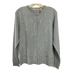 May include: A light blue cardigan sweater with long sleeves and a button-up front. The sweater features delicate embroidered leaf and floral designs with small embellishments. The brand tag reads "CAROLYN TAYLOR".