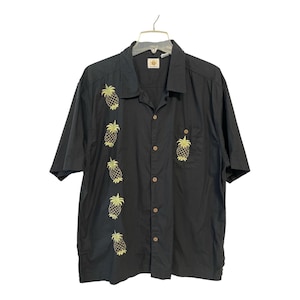 Può includere: Camicia nera a maniche corte con ananas gialli ricamati. La camicia ha una tasca sul petto e bottoni in legno. La camicia è appesa a una gruccia su sfondo bianco.