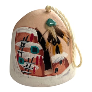 May include: A hand-painted ceramic ornament with a Native American design. The ornament features a peach-colored base with a white border and painted details in red, black, and turquoise. It is adorned with feathers and a turquoise stone, and has a cream-colored string for hanging.
