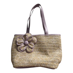 May include: A woven straw tote bag with a large flower applique. The bag features lavender-colored trim and handles. The flower is a matching lavender color with a button center. The bag is rectangular in shape.