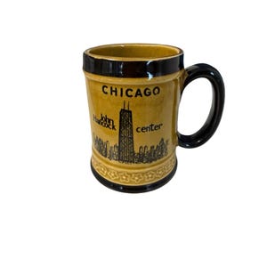 May include: A tan ceramic mug with a black handle and rim. The mug features the word "CHICAGO" above a black illustration of the John Hancock Center and Chicago skyline. The mug has a decorative floral pattern at the base.