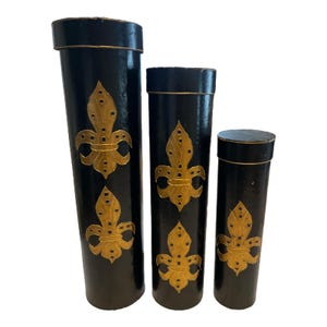 May include: A set of three black cylindrical boxes, each adorned with a gold fleur-de-lis motif. The boxes are of varying heights, with the tallest measuring around 30 cm. Gold trim accents the lids of each box.