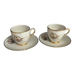 May include: Two matching cream-colored teacups and saucers. Each cup and saucer has a gold rim and a decorative emblem with red, green, and gold accents. The cups have a looped handle.