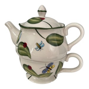 May include: A white ceramic teapot and teacup set. The teapot has a lid and spout, and the teacup is stacked underneath. The set is decorated with green leaves, ladybugs, and butterflies. The teapot has a handle and a small knob on the lid.