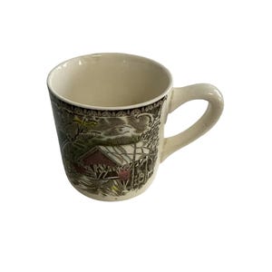 Könnte beinhalten: Ein Keramikbecher mit cremefarbener Innenseite und einem Henkel auf der rechten Seite. Die Außenseite zeigt eine detaillierte, mehrfarbige Illustration einer überdachten Brücke in einer Winterlandschaft mit Bäumen und Schnee.