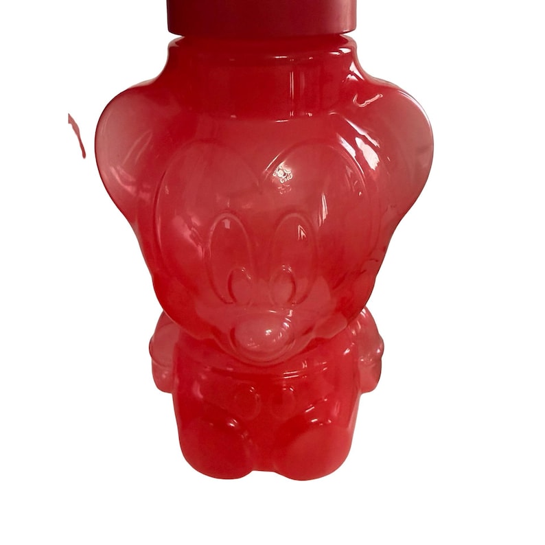 May include: A translucent red plastic bottle shaped like the character Mickey Mouse. The bottle features a molded relief of the character's face and body, including large rounded ears and a circular cap at the top. The material is semi-transparent.