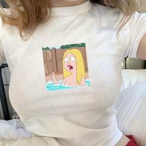 Op de afbeelding: Wit T-shirt met een cartoonafbeelding van een blonde vrouw in een zwembad, die een sigaret rookt. De cartoon staat in een rechthoekige lijst. Het T-shirt is gemaakt van een zacht, lichtgekleurd materiaal.