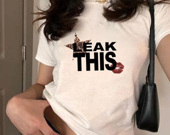 May include: White short-sleeved t-shirt with the text "LEAK THIS" in black block letters. A leopard print star and red lips graphic are also featured. The shirt is a casual style.