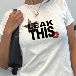 May include: White t-shirt featuring the text "LEAK THIS" in black. A star with a leopard print pattern is above "LEAK", and a red lipstick kiss graphic is beside "THIS".