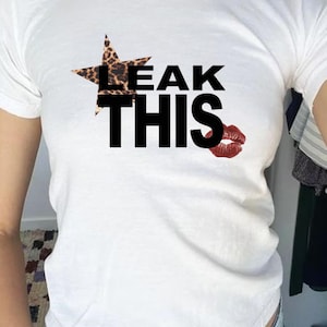 May include: White t-shirt featuring the phrase "LEAK THIS" in black, bold font. A star with a leopard print pattern is above "LEAK". A red lipstick kiss is printed to the right of the text.