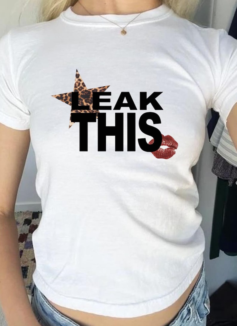 May include: White t-shirt featuring the phrase "LEAK THIS" in black, bold font. A star with a leopard print pattern is above "LEAK". A red lipstick kiss is printed to the right of the text.