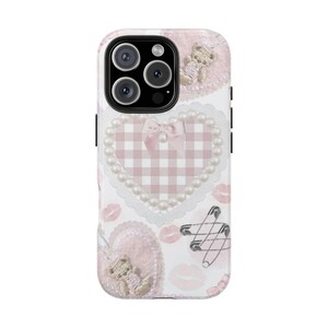 Peut inclure: Coque de téléphone blanche ornée de cœurs roses, d'ours en peluche et d'empreintes de lèvres. La coque présente un cœur à carreaux roses avec une bordure de perles et un nœud. Des épingles de sûreté sont également incluses.