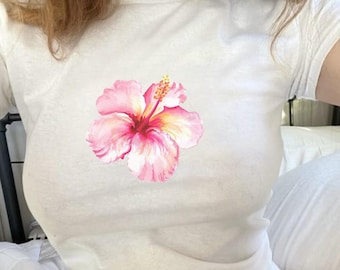 May include: White t-shirt featuring a large pink and yellow hibiscus flower graphic. The flower has detailed shading and a prominent stamen. The shirt is a crew neck style and appears to be made of a soft, lightweight material.