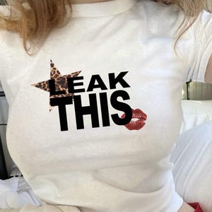May include: White t-shirt featuring the text "LEAK THIS" in black. The word "LEAK" is partially covered by a star-shaped leopard print design. A red lipstick kiss graphic is next to the word "THIS".