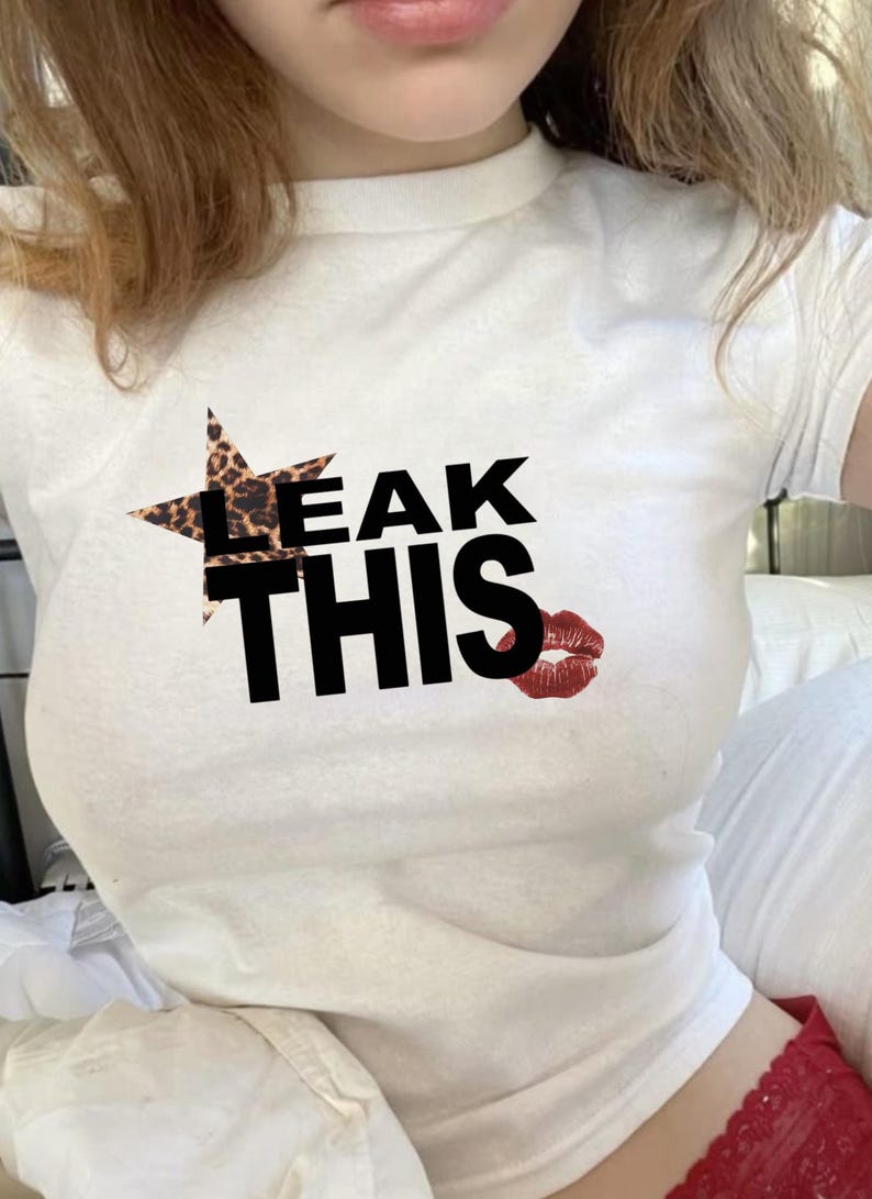 May include: White t-shirt featuring the text "LEAK THIS" in black. The word "LEAK" is partially covered by a star-shaped leopard print design. A red lipstick kiss graphic is next to the word "THIS".