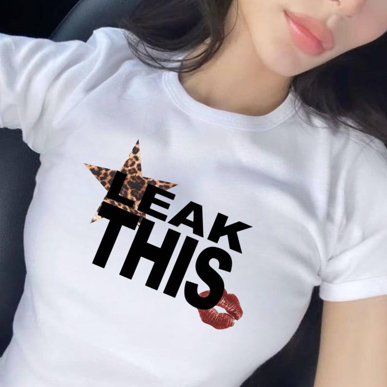 May include: White t-shirt featuring the text "LEAK THIS" in black, a leopard print star, and a red lip graphic. The top has short sleeves and a crew neck.