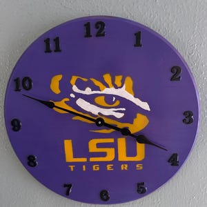 May include: A round purple clock with black hands and numerals. The clock face features the LSU Tigers logo in yellow and white, with the text "LSU TIGERS" in yellow. The clock is mounted on a light gray wall.