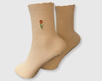 May include: A pair of beige ribbed socks with a scalloped cuff. One sock features an embroidered tulip in shades of orange, green, and brown. The soles have small, dark gray non-slip grips. These socks are designed for comfort and style.