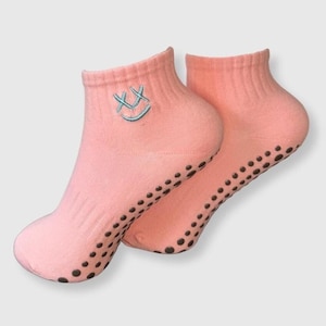 May include: A pair of light pink ankle socks with a teal embroidered smiley face. The soles have small, dark brown, non-slip grips. These socks are designed for comfort and grip.