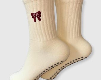 May include: A pair of cream-colored socks with a burgundy bow embroidered on the cuff. The socks have a ribbed texture and small, dark gray non-slip dots on the soles. These socks are designed for comfort and style.