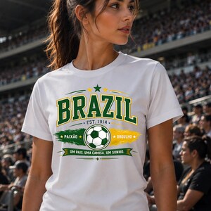 May include: A white short-sleeve t-shirt featuring a graphic design with the word BRAZIL in green letters, a soccer ball, and the text EST. 1914, PAIX&Atilde;O, ORGULHO, and UM PAIS. UMA CAMISA. UM SONHO. The design incorporates green and yellow brush strokes.
