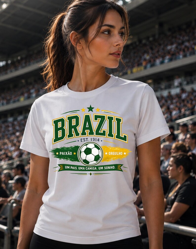 May include: A white short-sleeve t-shirt featuring a graphic design with the word BRAZIL in green letters, a soccer ball, and the text EST. 1914, PAIX&Atilde;O, ORGULHO, and UM PAIS. UMA CAMISA. UM SONHO. The design incorporates green and yellow brush strokes.