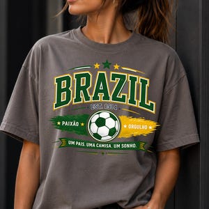 May include: A charcoal gray t-shirt featuring a graphic with the word BRAZIL in green and white letters, a soccer ball, the text EST. 1914, PAIX&Atilde;O, ORGULHO, and the phrase UM PAIS. UMA CAMISA. UM SONHO. The design includes green and yellow brush strokes.