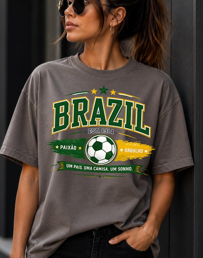 May include: A charcoal gray t-shirt featuring a graphic with the word BRAZIL in green and white letters, a soccer ball, the text EST. 1914, PAIX&Atilde;O, ORGULHO, and the phrase UM PAIS. UMA CAMISA. UM SONHO. The design includes green and yellow brush strokes.