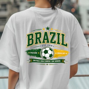 May include: A white t-shirt featuring a graphic on the back with the word BRAZIL in large green letters, the text EST. 1914, a soccer ball, the words PAIX&Atilde;O and ORGULHO, and the phrase UM PAIS. UMA CAMISA. UM SONHO. in green, yellow, and white colors.