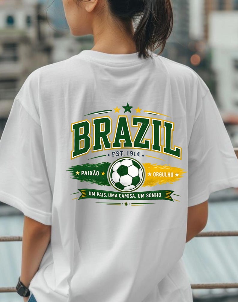 May include: A white t-shirt featuring a graphic on the back with the word BRAZIL in large green letters, the text EST. 1914, a soccer ball, the words PAIX&Atilde;O and ORGULHO, and the phrase UM PAIS. UMA CAMISA. UM SONHO. in green, yellow, and white colors.